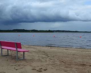 Eine pinke Bank am Strand mit Blick auf den See und Wolken.